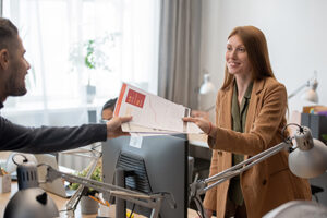 Young smiling businesswoman passing financial papers to male colleague over computer monitor