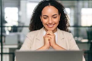 Satisfied brazilian or hispanic curly haired business woman, company ceo, recruitment manager, sits at a work desk in a modern creative office, working in a laptop, looks at screen, smiling friendly